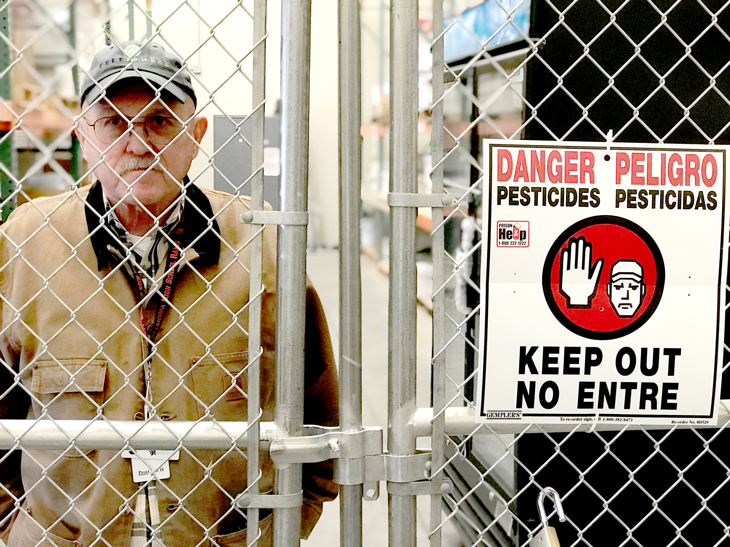 Dan Vezie behind a fence. The fence has a sign on it saying "Danger, Pesticides, Keep Out" in English and Spanish.