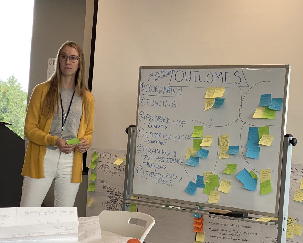 Annie Krueger of Compliance Services International stands next to a whiteboard titled "Outcomes." There are both whiteboard marker notes and sticky notes on the whiteboard.