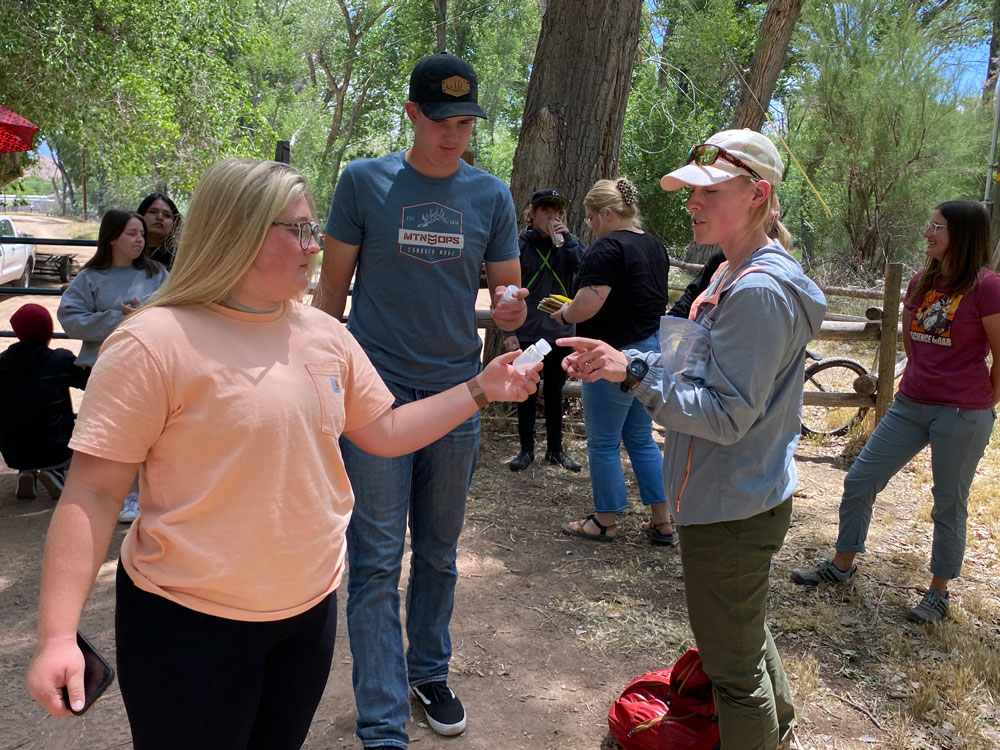 Michele Rehbein hands small plastic containers to Harlie Book and Lane Berry in front of a natural area.