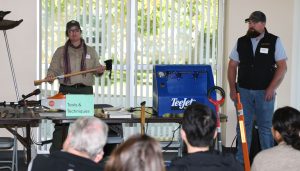 Rachel Kesel and Tom Getts stand by a table full of weed management tools. Rachel holds an axe.